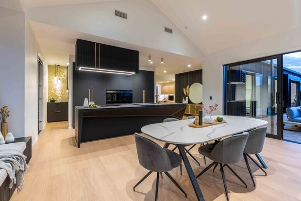 Open-plan contemporary kitchen and dining area with a large black island, integrated dark cabinetry, pale timber flooring, and a round marble-look dining table with upholstered chairs. Floor-to-ceiling black-framed glazing opens toward an outdoor area, while warm recessed lighting and decorative accents create a refined, inviting interior.