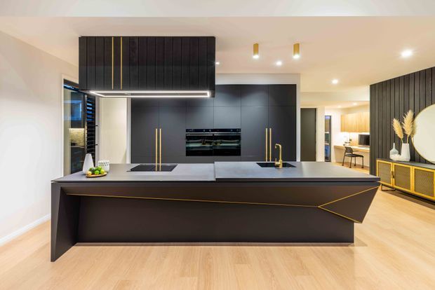 Contemporary open-plan kitchen dominated by a large dark island with a sculptural angular front, integrated black cabinetry, brass-gold accents and a suspended dark rangehood. Pale timber flooring, warm white walls, recessed lighting and a glimpse of an adjacent dining or living area create a refined, high-contrast interior.