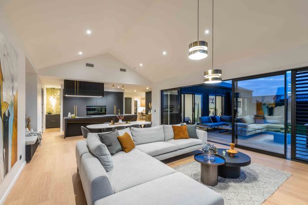 Contemporary open-plan living space with a large light-grey sectional sofa, round coffee tables, pendant lighting and a neutral textured rug. The living area connects directly to a sleek dark kitchen with an island, while expansive black-framed glazing opens toward an outdoor pool terrace. Light timber flooring, white walls and warm ambient lighting create an inviting, polished interior.