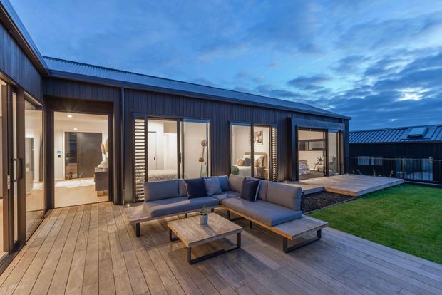 Contemporary single-storey home exterior viewed from a spacious timber deck at dusk. The dark vertically clad facade has extensive sliding glass doors opening onto an outdoor lounge area with sectional seating and a low coffee table. Interior living spaces are visible through the glazing, with a neatly landscaped lawn and a separate dark-roofed structure beyond.