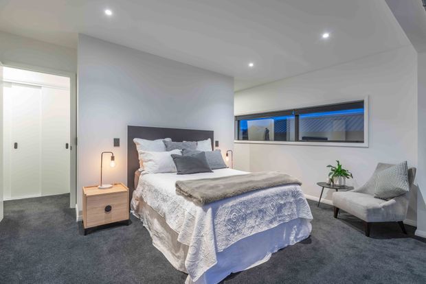 A contemporary bedroom with a neatly made bed, upholstered headboard, bedside table and lamp, grey carpet, recessed ceiling lights, built-in wardrobe doors, and a large horizontal window. The room uses a soft neutral palette with blue-grey bedding and a compact upholstered armchair.