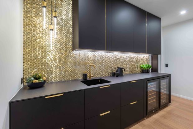 A sophisticated contemporary kitchen with matte charcoal cabinetry, slim brass pulls and accents, a dark benchtop, integrated black sink and brass tap. A gold-toned textured mosaic backsplash extends to the ceiling behind the cabinetry, complemented by pendant lights and under-cabinet lighting. Glazed beverage or wine-storage cabinets sit at the right, with warm timber flooring and minimal white walls.