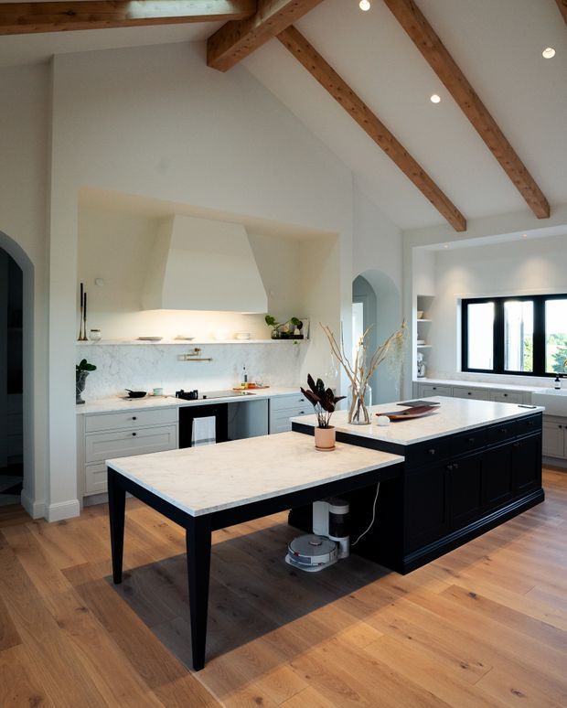 Bright, spacious kitchen with a large central island and separate light-toned worktable, dark painted cabinetry, pale stone benchtops, white walls, and exposed timber roof beams. The room features a pitched ceiling, recessed lighting, wide timber flooring, arched openings, and black-framed windows.