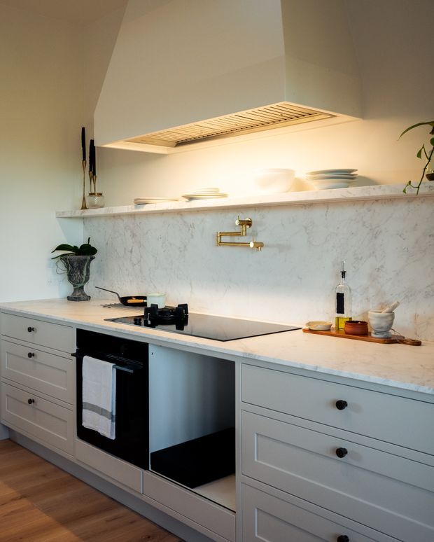 A refined, light-toned kitchen with pale grey shaker-style cabinetry, a veined marble backsplash and benchtop, integrated black appliances, a flush induction cooktop, and a brass wall-mounted pot filler. A sculptural white rangehood with a timber slatted underside dominates the upper wall, while warm timber flooring and restrained decorative pieces add warmth.
