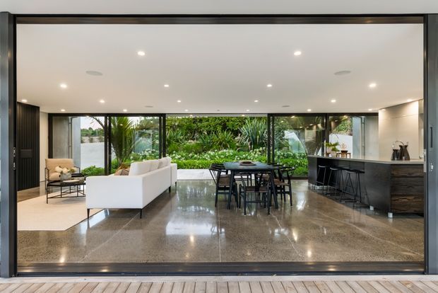 A refined open-plan interior viewed from a timber deck through fully glazed sliding doors. The space combines a living area, dining table and kitchen with a dark island and cabinetry. Polished speckled stone or concrete flooring, dark framed glazing, recessed ceiling lights and extensive garden-facing glass create a bright, indoor-outdoor connection.