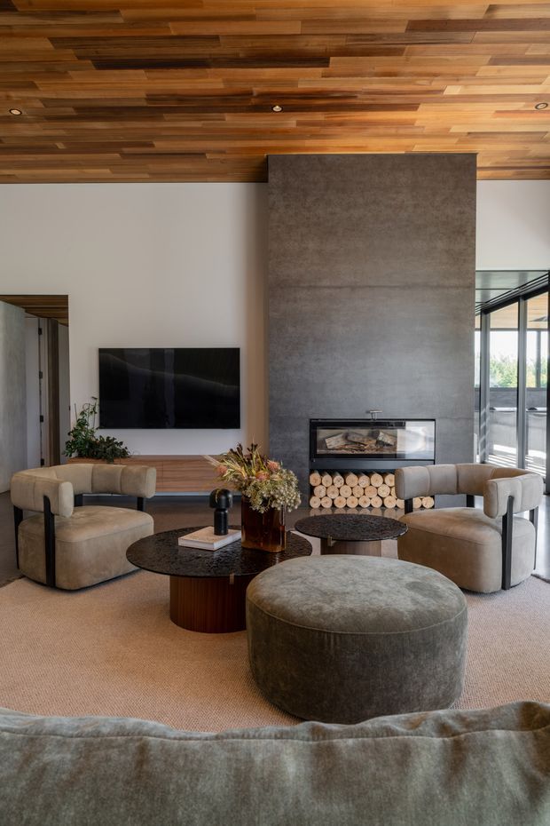 A sophisticated living room featuring a tall concrete fireplace surround with integrated firebox and timber log storage, a wall-mounted television, and extensive warm timber lining across the ceiling. Two rounded upholstered armchairs, a large circular ottoman, and dark round coffee tables form a conversational seating arrangement over a neutral carpet. Full-height glazing on the right opens the room to the outdoors, while earthy upholstery, timber, concrete, and muted neutral tones create a refined contemporary atmosphere.