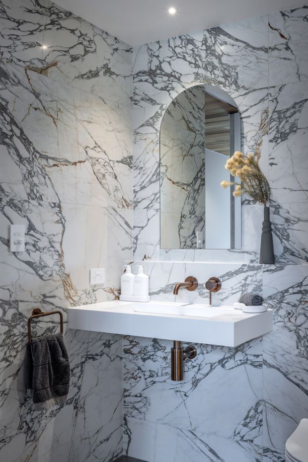 A compact, highly finished bathroom with large-format white marble-look wall and floor tiles featuring dramatic grey and warm brown veining. A floating white vanity with integrated basin is paired with exposed brushed bronze/copper-toned wall-mounted tapware and matching plumbing. An arched mirror is set into the tiled wall, with a dark towel rail, charcoal towel, minimalist black vase, and recessed ceiling lighting.