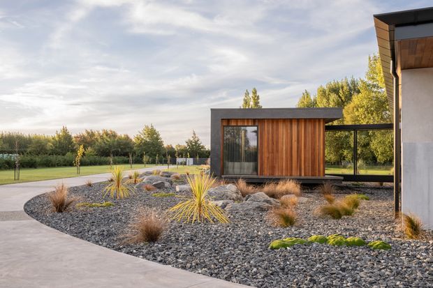 Contemporary single-storey architectural exterior set within a landscaped garden. The image shows a compact pavilion-like structure with dark charcoal cladding and vertical warm timber wall panels, extensive glazing, and a flat roof. A second building volume is partially visible at right, connected by a dark-framed glazed passage. The foreground features a curved concrete path and a low-maintenance gravel garden with ornamental grasses, sculptural rocks, and young trees beyond.