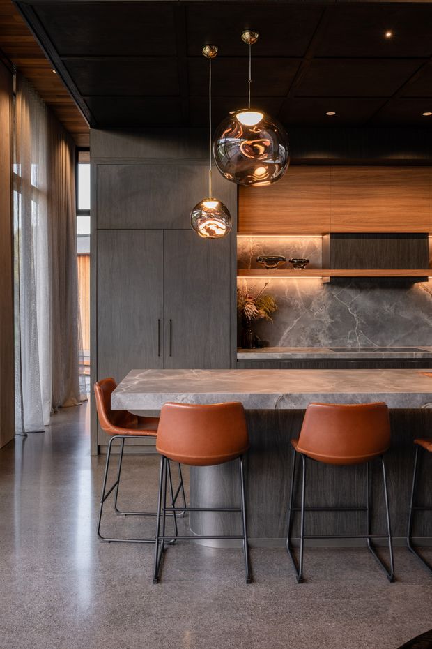 A sophisticated contemporary kitchen centered on a large island with a pale veined stone benchtop and dark timber cabinetry. Three brown leather bar stools line the island, while reflective spherical pendant lights hang above. The kitchen features a dark coffered or panelled ceiling, tall integrated cabinetry, warm timber wall cabinetry, open shelving with under-shelf lighting, and a dramatic dark marble-look splashback. Full-height sheer curtains and polished concrete flooring contribute to the refined, moody atmosphere.