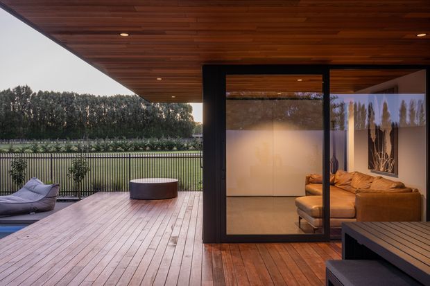 A covered timber deck and outdoor living area adjoining a glazed pavilion or living room. The space features warm timber ceiling and decking, slim dark-framed glazing, built-in recessed lighting, an outdoor lounge chair, low round table, and a partially visible indoor sectional sofa. The deck overlooks a fenced landscaped garden and lawn, creating a calm indoor-outdoor connection.