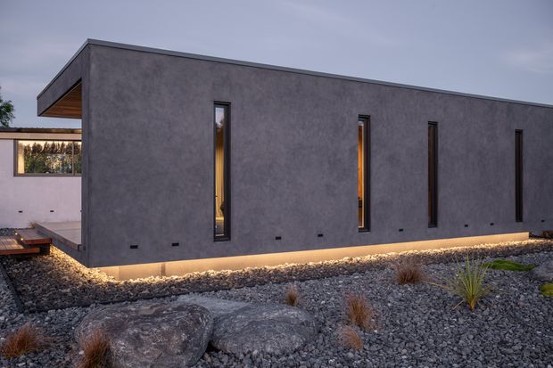 Exterior view of a long, minimalist rectangular building with a dark grey textured rendered facade, narrow vertical windows, a flat roof, and warm linear lighting at the base. The building is set within a gravel xeriscaped landscape with large rocks and sparse ornamental grasses, with a partially visible adjoining structure and timber-lined soffit at left.