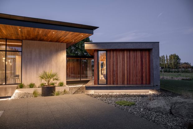 Contemporary low-profile residential exterior at dusk, featuring a projecting flat roof with a warm timber-lined soffit, dark rendered walls, extensive glazing, and a compact timber-clad garage or outbuilding. The paved approach and gravel landscaping frame the modern architectural composition.