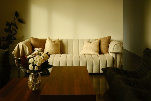 Warm, softly lit living room with a cream channel-tufted sofa, textured neutral cushions, dark lounge seating, and a rich timber coffee table. A floral arrangement and leafy plant add organic accents, while the low golden lighting creates an intimate, refined atmosphere.