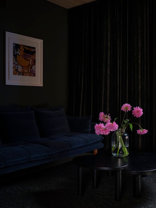 A dark, moody living room featuring a deep navy velvet sofa, black floor-to-ceiling pleated curtains, a round black coffee table, dark flooring, and a glass vase of vivid pink flowers. A framed artwork hangs on the dark wall, while a timber-toned ceiling section adds subtle warmth.