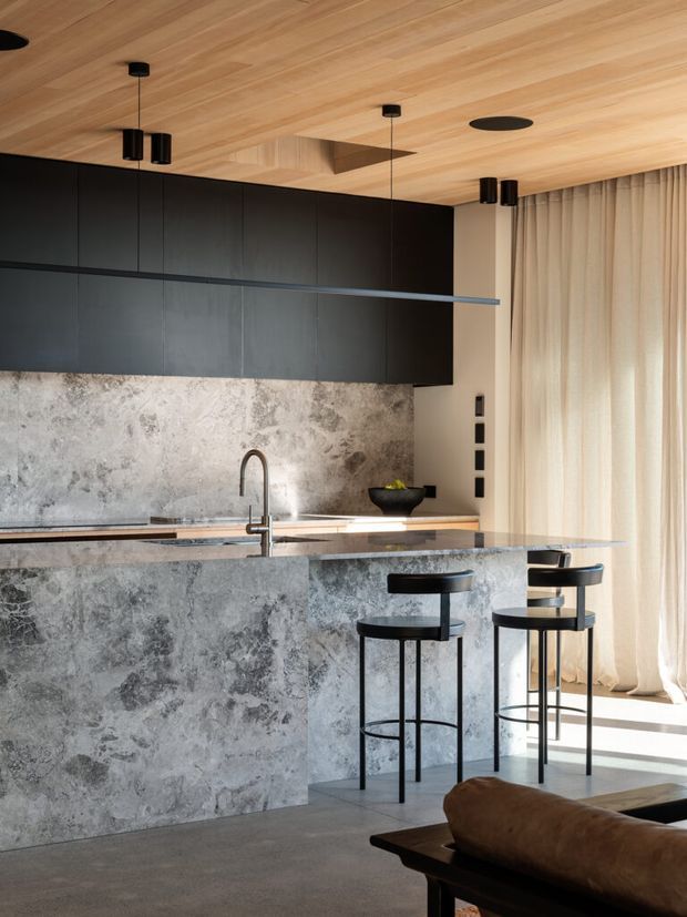 A refined contemporary kitchen with a large stone-clad island and matching splashback, matte-black overhead cabinetry, integrated black pendant and recessed lighting, and a timber-lined ceiling. Two black bar stools sit at the island, while full-height sheer curtains bring soft daylight into the space. A partial leather sofa is visible in the foreground, suggesting an adjoining living area.