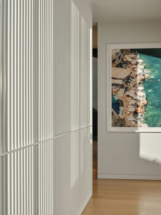 A bright, minimalist interior hallway or entry passage featuring a full-height white vertical fluted/slatted partition wall. Warm timber flooring continues through the space, with a framed coastal photograph on a pale neutral wall and strong sunlight casting linear shadows across the surfaces.