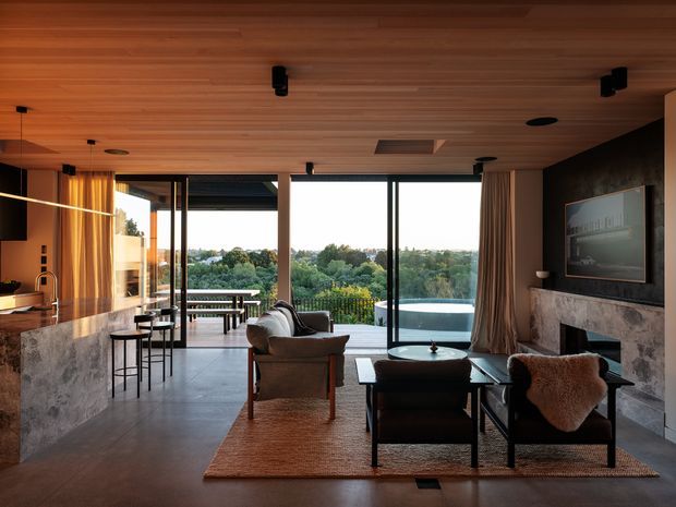 A refined open-plan living, dining and kitchen interior with floor-to-ceiling glazing opening to a terrace and pool. The room features a warm timber-lined ceiling, dark-framed glass doors, stone kitchen island, dark textured fireplace wall, built-in fireplace, contemporary lounge furniture, and a panoramic leafy outlook.