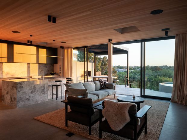 A warm, open-plan contemporary living and kitchen space with extensive floor-to-ceiling glazing opening to a balcony and landscaped view. The room features a timber-lined ceiling, dark flooring, a sculptural stone kitchen island with integrated seating, timber and dark cabinetry, a neutral sectional sofa, leather armchairs, and soft layered textiles. Recessed and pendant lighting create an intimate atmosphere, while a glimpse of a circular outdoor spa or pool is visible beyond the glazing.