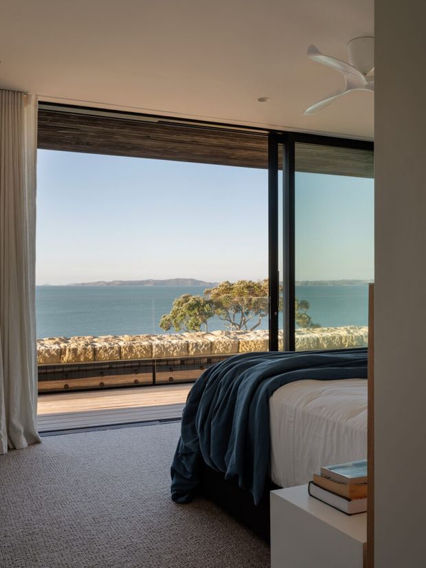 A serene modern bedroom opening onto a waterfront balcony or terrace. The room features a low-profile bed with dark blue bedding, pale carpet, white curtains, a white bedside table with books, and expansive black-framed glazing. A timber roller blind and a ceiling fan are visible, with the ocean and rocky coastline beyond.