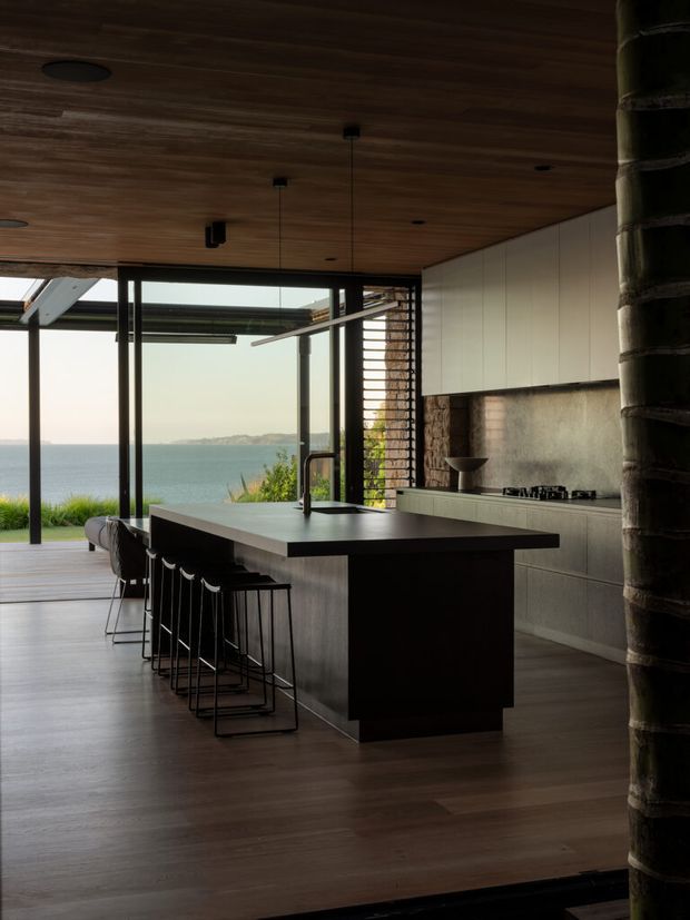 A dark, refined contemporary kitchen opening directly onto a waterfront view. The room features a large central island with an integrated sink and seating for several people, full-height glazing with slim dark frames, warm timber ceiling and flooring, pale built-in cabinetry, and a textured stone or concrete-look splashback. A substantial round structural column frames the right side of the image.