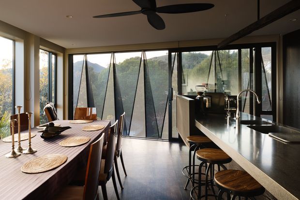 A contemporary open-plan kitchen and dining area with a long dark dining table, timber dining chairs, woven placemats, and decorative brass candlesticks. The kitchen features a large dark stone island with an integrated sink, curved faucet, black stools with timber seats, dark cabinetry, and extensive floor-to-ceiling glazing. Angled external sun-shading panels are visible through the windows, with leafy hills beyond. A large black ceiling fan and exposed dark beam add strong architectural character.