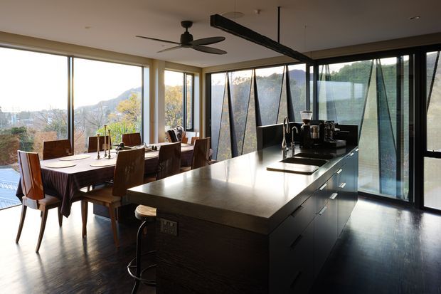 A modern open-plan kitchen and dining area with a large dark island, integrated cooktop and sink, dark cabinetry, and a timber dining table with upholstered chairs. Full-height glazing wraps the room, providing expansive views of a wooded, hilly landscape. A sculptural black ceiling fan and linear ceiling feature add contemporary detail.