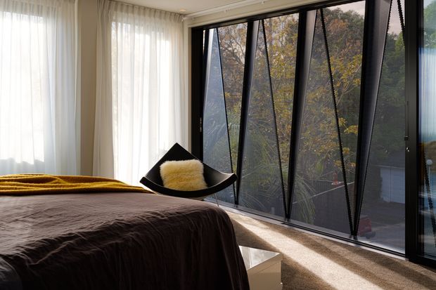 A contemporary bedroom with a low platform bed dressed in dark brown bedding and a mustard throw, positioned beside expansive black-framed glazed windows. Sheer white curtains soften the daylight, while a sculptural black lounge chair with a cream cushion sits near the glazing. The room has warm carpet flooring and a low white bedside table, with trees visible outside.