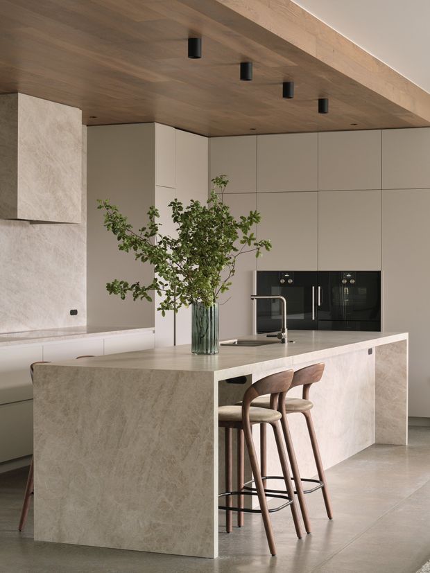 A refined contemporary kitchen featuring a large pale stone island with waterfall edges, integrated taupe cabinetry, black built-in appliances, and a warm timber-lined ceiling with recessed black spotlights. Two sculptural timber bar stools sit at the island, while a glass vase with leafy branches adds a natural accent.