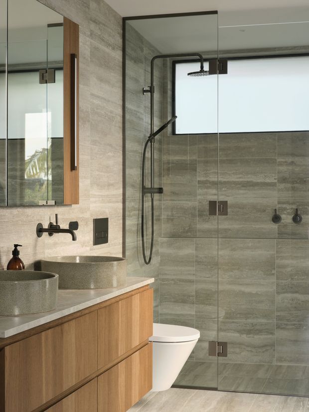 Contemporary bathroom featuring a walk-in glass shower with dark metal hardware, large grey stone-look wall and floor tiles, a timber vanity with integrated basin, vessel basin, wall-mounted black tapware, mirrored cabinetry, and a partially visible toilet. Natural timber and muted grey finishes create a refined, modern atmosphere.