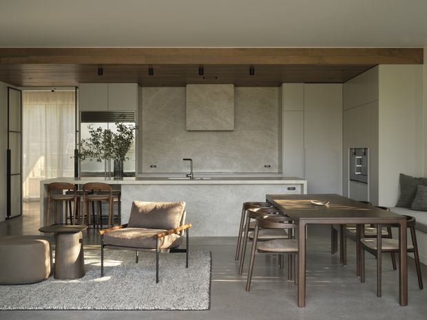 Open-plan contemporary kitchen and dining/living area with a long central island, pale integrated cabinetry, a textured light stone or plaster splashback, warm timber ceiling lining and beam, and floor-to-ceiling glazing at the left. A dark timber dining table and chairs occupy the foreground, with a lounge chair, ottomans and rug creating a relaxed seating zone.