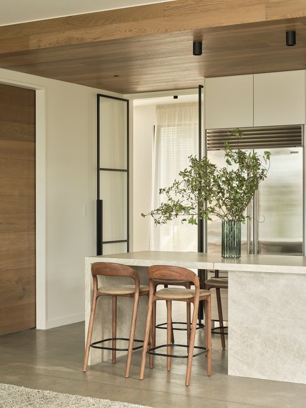 A refined contemporary kitchen with a large pale stone island and integrated breakfast bar, paired with curved timber bar stools. The space features warm timber ceiling panelling and wall cabinetry, soft off-white surfaces, black-framed glazed internal doors, stainless-steel refrigeration, and a polished concrete-look floor. A tall glass vase with leafy branches adds a natural focal point.