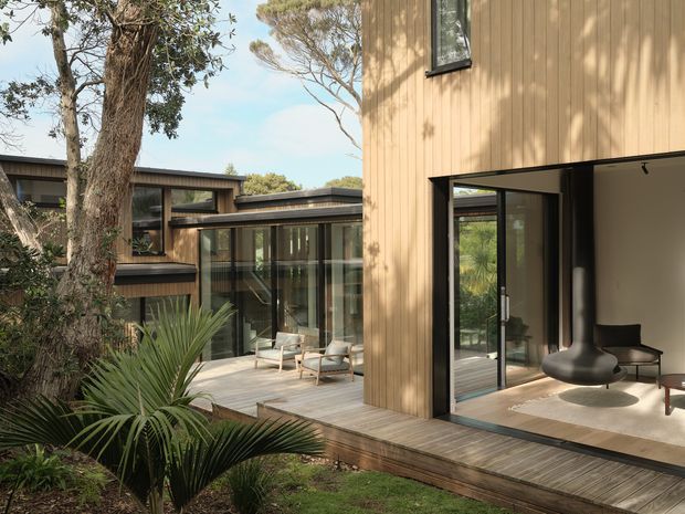 A contemporary timber-clad house arranged around a landscaped courtyard, viewed from the garden. The image shows warm vertical wood cladding, extensive dark-framed glazing, a timber deck, and open-plan interior spaces with minimalist furniture visible through the open doors. Mature trees and palm-like planting surround the building.