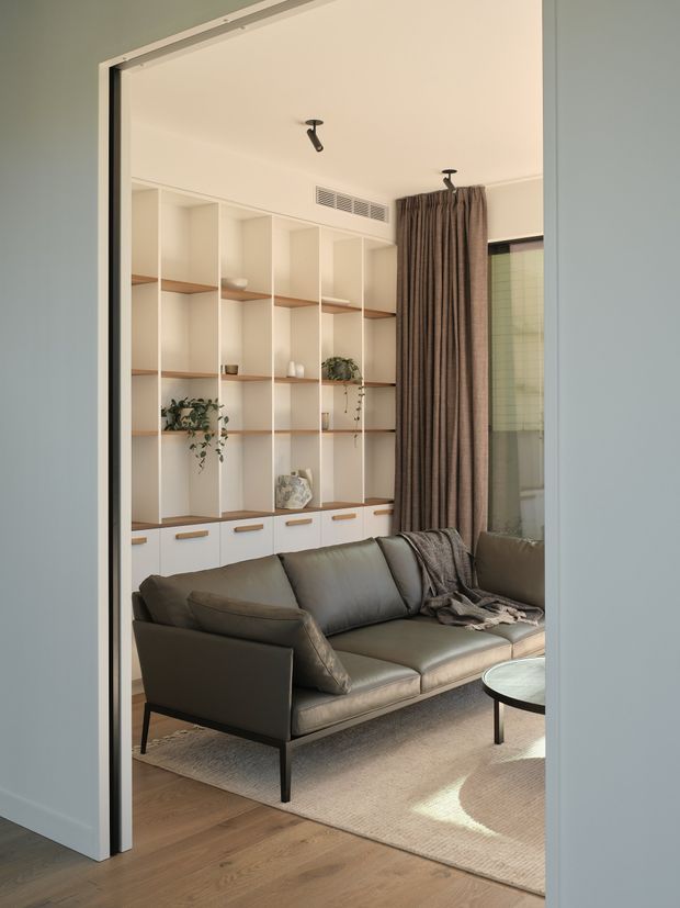 A softly lit contemporary living room viewed through a wide doorway. The room features a low-profile dark taupe leather sofa and armchair, a round coffee table, a large neutral rug, warm timber flooring, and full-height built-in shelving with white cabinetry, open shelves, and wood-toned shelf inserts. Floor-to-ceiling brown drapery frames a window, while minimalist black track spotlights and a linear air vent complete the clean interior.
