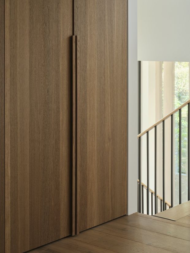 Close-up interior view of a tall, full-height timber-veneered built-in cupboard or wardrobe with long vertical timber handles. The warm wood cabinetry stands beside a stairwell with slim black metal balustrades and a timber handrail, with natural light entering through large glazing.