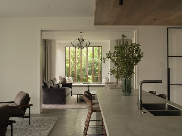 A refined open-plan kitchen and living area viewed from the kitchen island. The foreground features a large grey stone island with an undermount sink, dark tapware and timber bar stools. Beyond it is a calm living room with dark sofas, a pale rug, a built-in window seat and full-height timber-framed glazing overlooking greenery. A decorative chandelier, indoor tree and warm timber ceiling lining add natural character to the light neutral interior.