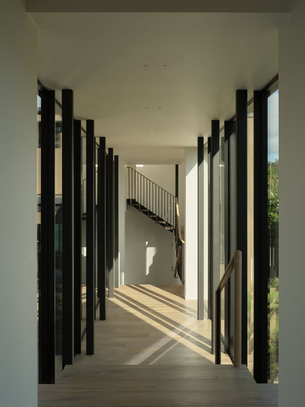 A long, bright interior corridor with pale timber flooring, white walls and ceiling, and extensive full-height glazing framed in black. Strong sunlight creates elongated shadows across the floor. Black vertical structural or screening elements line both sides, leading toward a minimalist open staircase with a slim dark metal balustrade at the far end.