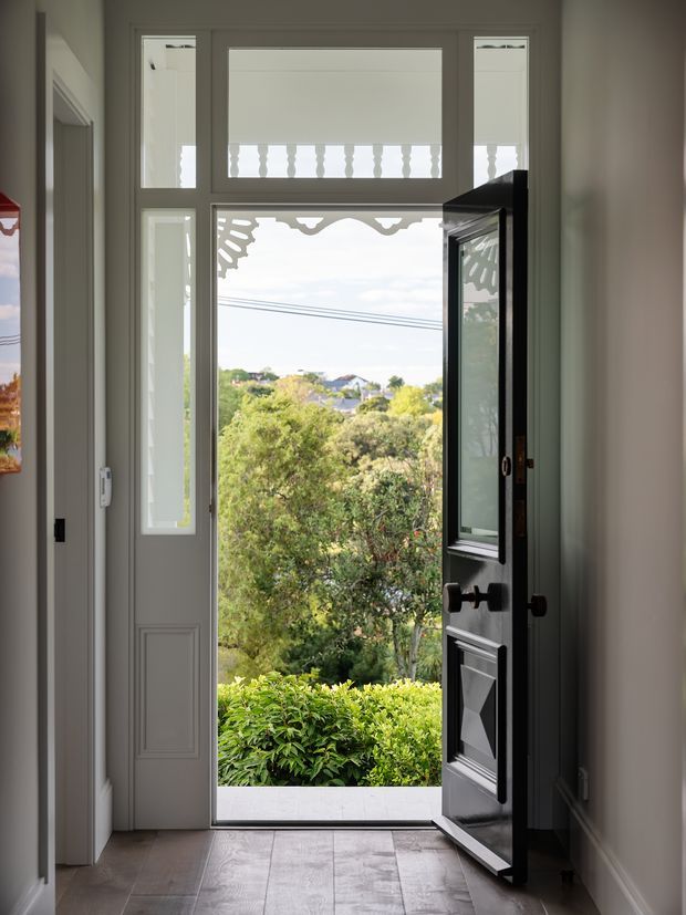 Interior view of a refined residential entryway with a tall, dark-painted glazed front door standing open. Decorative upper glazing and sidelights bring in daylight, while patterned tile flooring and pale walls frame a leafy suburban garden and distant rooftops beyond the entrance.