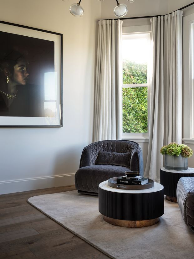Refined living room corner with pale walls, tall curtained sash windows, dark timber flooring, a soft neutral rug, quilted charcoal lounge chairs, and a round black ribbed coffee table with a timber base. A large framed portrait and fresh floral arrangement add decorative detail, while contemporary ceiling spotlights provide focused illumination.