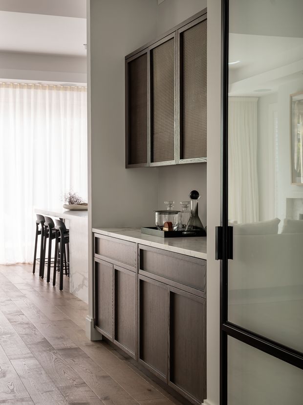 A refined built-in drinks cabinet and storage wall in a contemporary home. Dark timber cabinetry with textured mesh-front upper doors and drawers sits against a warm grey wall, with a pale stone benchtop and styled glassware. A black-framed glazed door reflects a bright living area, while the adjacent dining or kitchen zone has a bar-height island, stools, sheer curtains, and pale timber flooring.