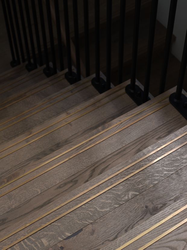 Close-up view of a contemporary staircase with wide, weathered-looking timber treads featuring narrow brass or gold-toned inlay strips. Slim black metal vertical balusters and dark metal mounting brackets form the handrail or balustrade, creating a strong linear pattern. The image is dimly lit and focused on the stair construction and material detailing.