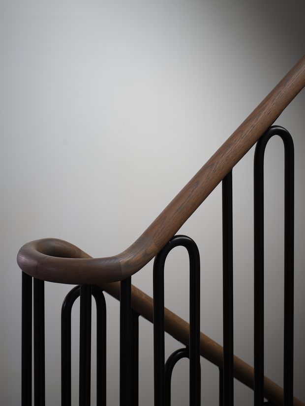 Close-up architectural detail of a staircase balustrade, featuring a sculpted dark timber handrail with a curved transition and slender black metal vertical loop-like balusters against a plain light wall.