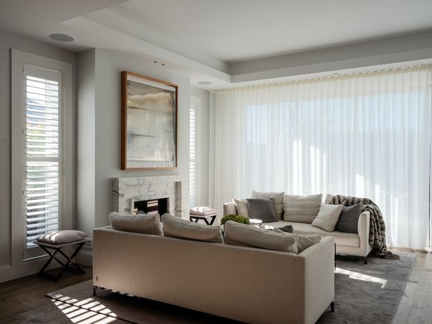 Bright, refined living room with a large neutral sectional sofa arranged around a marble-surround fireplace. Full-height sheer curtains, plantation shutters, layered neutral furnishings, a framed artwork, recessed ceiling details, and a soft area rug create an airy contemporary-classic interior.