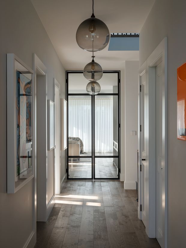 A long, refined residential hallway with pale walls, white panelled doorways, and a grey-brown timber floor. Three spherical pendant lights hang along the central axis, while black-framed glazed doors at the end reveal a bright bedroom or living area beyond. Framed artwork, a skylight, and strong daylight add visual interest to the otherwise restrained interior.