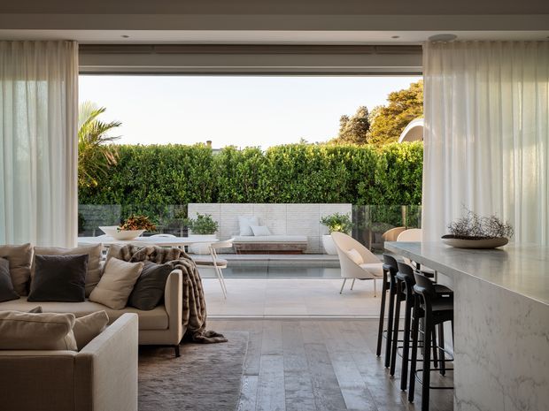 Open-plan contemporary living and dining space with a large fully retractable glazed opening leading to a landscaped outdoor terrace. The room features a neutral sectional sofa, pale timber or stone-look flooring, sheer curtains, a long marble or pale stone kitchen island with black bar stools, and a minimalist outdoor seating area beyond. The space is bright, refined, and strongly connected to the garden.