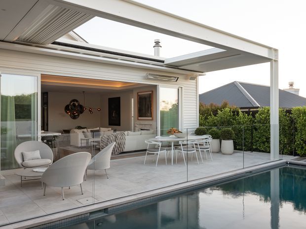 A contemporary indoor-outdoor living area opening directly onto a covered terrace and swimming pool. Large retractable roof panels and sliding glass doors create a seamless connection between the living room, dining area, terrace, and pool. The space features pale tiled flooring, white outdoor dining furniture, sculptural lounge chairs, glass pool fencing, and light horizontal exterior cladding. A neighbouring pitched-roof house and hedging are visible beyond the pool.