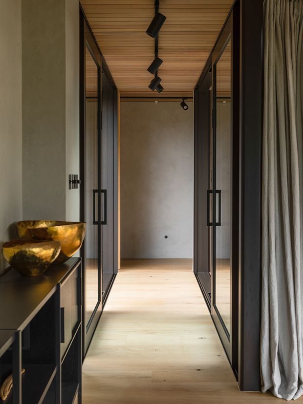 A narrow, refined interior hallway with pale timber flooring and a warm timber-lined ceiling. Full-height black-framed glass doors and partitions line both sides, creating a strong axial perspective toward a plain plaster wall. A dark built-in console with sculptural metallic bowls sits to the left, while full-height neutral curtains frame the right side. Black track lighting and dark hardware provide contrast against the warm, muted palette.