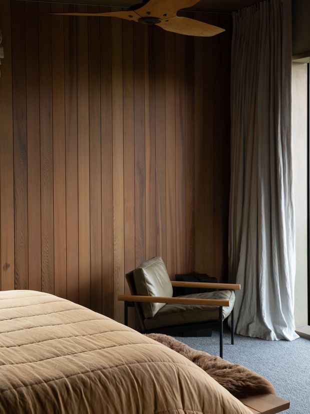A moody, refined bedroom with a full-height vertical timber-panelled feature wall, a low bed dressed in a warm beige quilt, and a leather lounge chair with wooden arms beside a dark side table. Floor-to-ceiling curtains frame a tall glazed opening, while a sculptural timber ceiling fan adds a natural focal point. The palette is rich in walnut tones, taupe, beige, and charcoal, with soft textured furnishings creating a calm atmosphere.