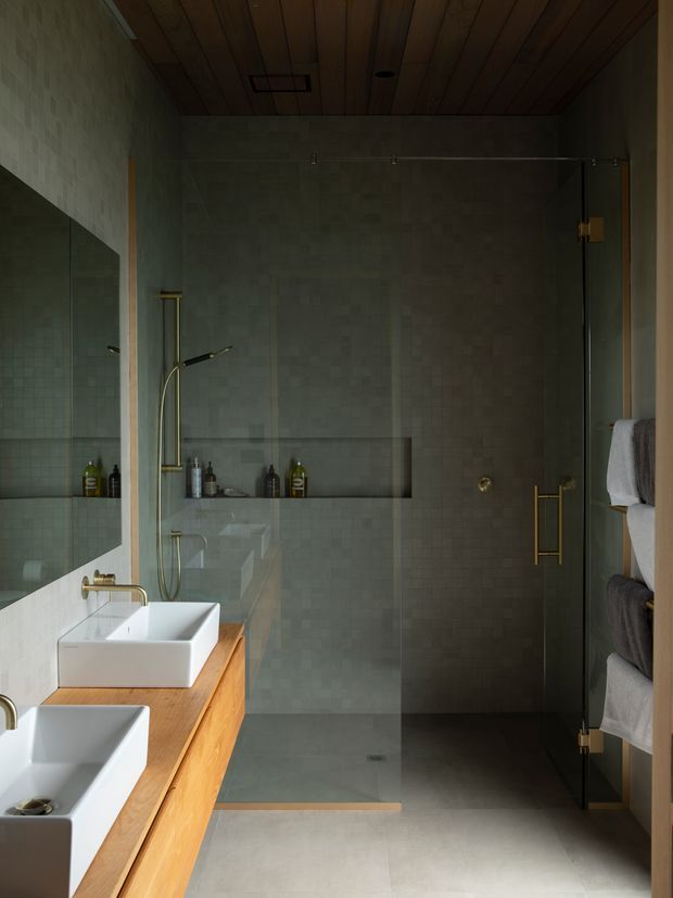 A moody, refined bathroom with a large frameless glass shower enclosure, pale small-format tiled walls, twin white vessel basins on a warm timber vanity, brass tapware and shower fittings, a timber-lined ceiling, and neatly arranged towels on open shelving.