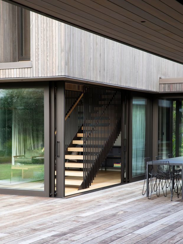 Contemporary timber-clad home viewed from a spacious timber deck, with large dark-framed glazed openings, an open stairway with slim vertical balustrade, and an adjacent dining area. The upper-level facade and soffit use linear timber boards, creating a clean, warm modern architectural composition.