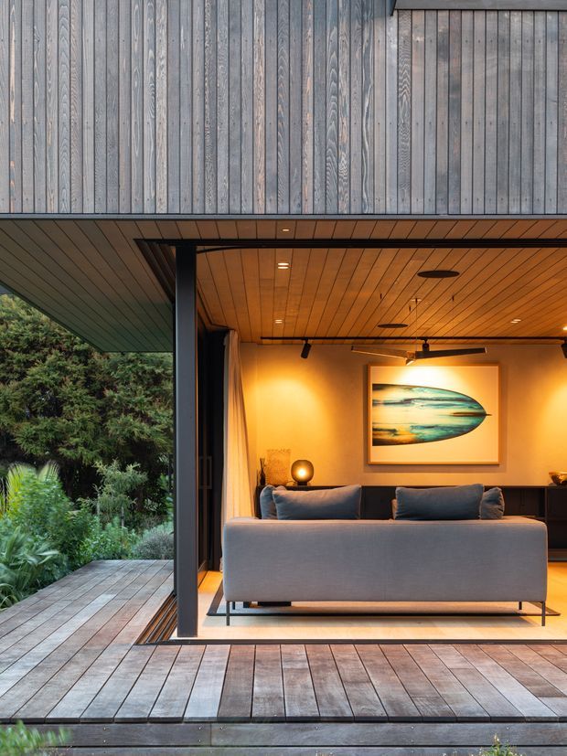 A contemporary indoor-outdoor living space opening onto a timber deck and lush garden. The room features a grey sofa, warm timber-lined ceiling and walls, dark structural framing, recessed and track lighting, and a framed abstract artwork. Vertical timber cladding forms the upper facade, while large open glazing or folding doors connect the living room to the deck.